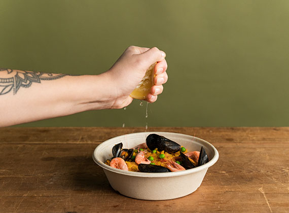 Hand squeezing lemon over seafood pasta contained in a BioPak round bowl with no added PFAS